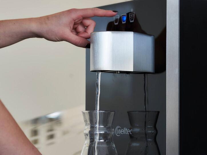 Eine Person füllt Wasser in eine Karaffe per Knopfdruck am Wasserspender