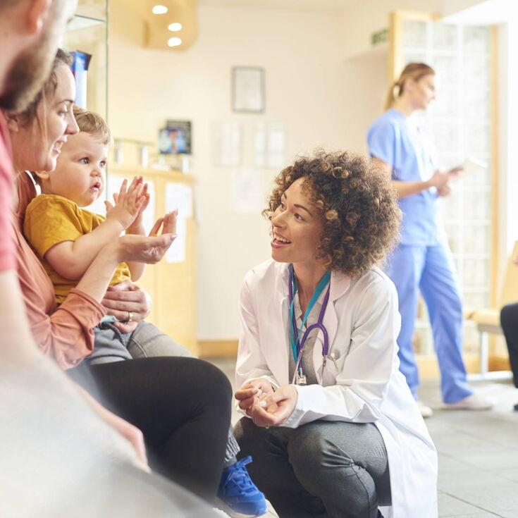 Im Wartezimmer eines Krankenhauses spricht eine Ärztin mit einem Kind, das bei seiner Mutter auf dem Schoß sitzt